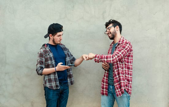 Two teenage friends greeting each other by fist bumping outdoors. Two smiling young guys bumping fists near a wall. Concept of two smiling friends bumping fists friendly