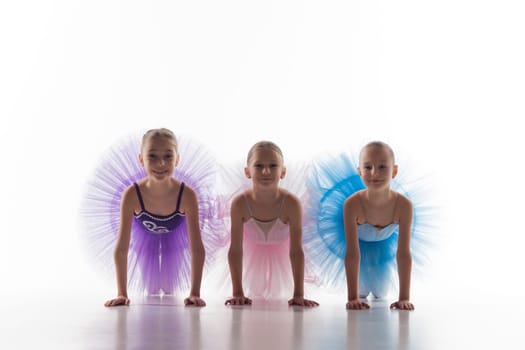 Three little ballet girls sitting in multicolored tutu and posing together on white background
