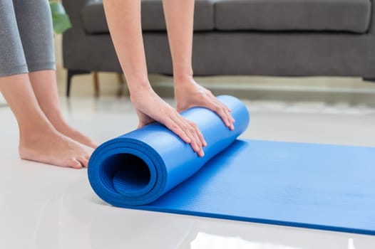 Cropped image of woman rolling blue mat after doing yoga exercise workout in living room at home. Healthy life and fitness training workout concept
