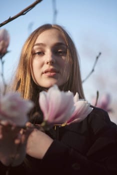 young blonde girl and pink blooming magnolia. happy girl standing near blooming tree in park. she has long straight hair and dark coat. magnolia flower near her face. she looks at camera