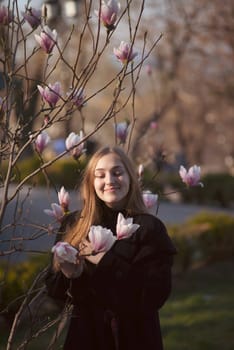 young blonde girl in the park near magnolia tree. happy girl laughing in the sun rays, pink flowers of the tree near her, she has long straight hair and dark coat