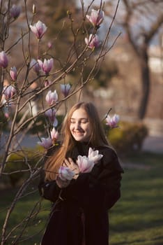 young blonde girl and blooming pink magnolia. happy girl laughing in the sun rays. she is standing near a blooming tree in the park. she has long straight hair and a dark coat.
