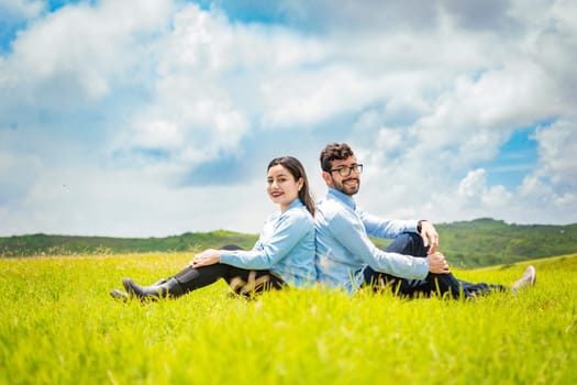 A wedding couple sitting with their backs to each other on the grass, Wedding couple in the field sitting with their backs to each other looking towards the camera