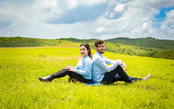 Couple in love in the field sitting back to back looking at the the camera, Young relaxed couple sitting in the field with their backs to each other lon the field