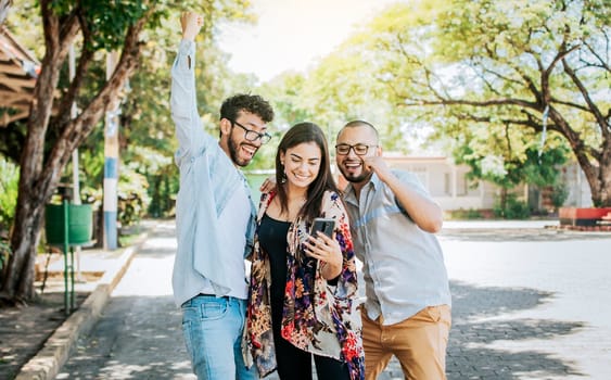 Three excited friends looking at the cell phone in the street. Three cheerful people looking at cell phone outdoors, Excited teenage friends looking at cell phone screen on the street