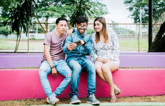 Three happy friends looking at the cell phone sitting in the park. Three cheerful people looking at cell phone sitting outside, happy teenage friends looking at phone screen outdoors
