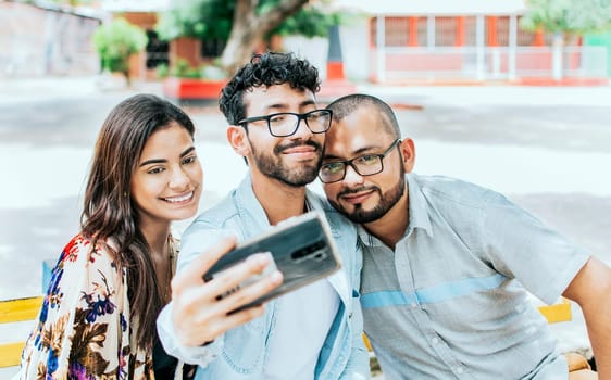 Three smiling friends sitting on a bench taking a selfie. Front view of three happy friends taking a selfie while sitting on a bench. Three friends taking a selfie on a park bench