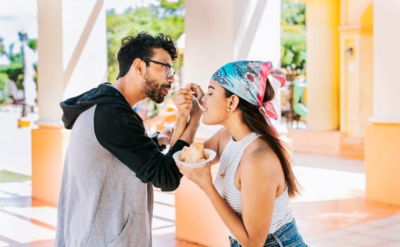 A couple eating shaved ice in a square, Young couple eating shaved ice together with arms intertwined, Side view of young couple with arms linked eating ice cream in a city square