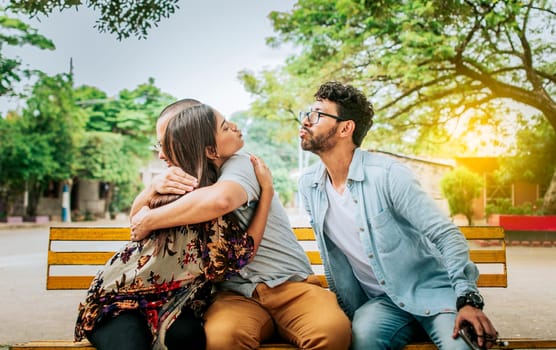 Unfaithful woman sitting hugging her boyfriend and secretly kissing another man. Unfaithful girlfriend hugging her boyfriend in a park secretly kissing another man, Couple infidelity concept