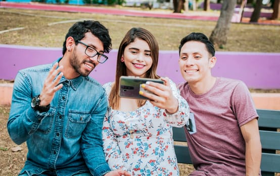 Three smiling friends taking a selfie sitting in the park, Three smiling friends sitting in the park taking a selfie. Front view of three happy friends taking a selfie while sitting on a bench