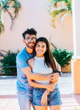 Portrait of a happy couple man hugging his girlfriend from behind, Portrait of a young couple man hugging his girlfriend from behind outside. Happy couple embracing looking at the camera