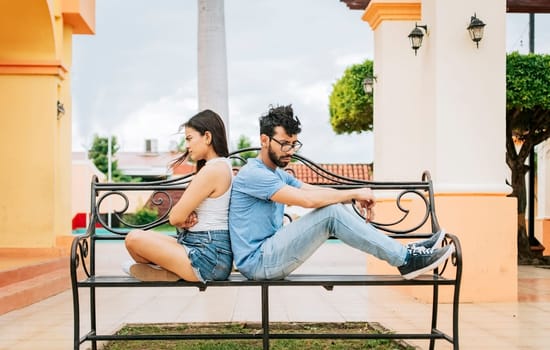Upset couple sitting back to back on a park bench. Man and girl arguing in a park. Upset young couple sitting back to back outside, young couple arguing sitting back to back