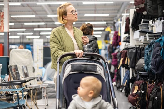 Casualy dressed mother choosing sporty shoes and clothes products in sports department of supermarket store with her infant baby boy child in stroller