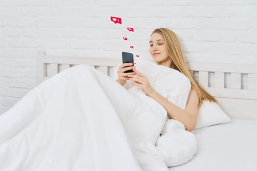 Young woman woke up and start browsing social media on mobile telephone while being in bed. Smiling woman likes photos in internet