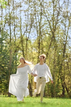 Happy newlyweds holding hands and running across the lawn in sunny day