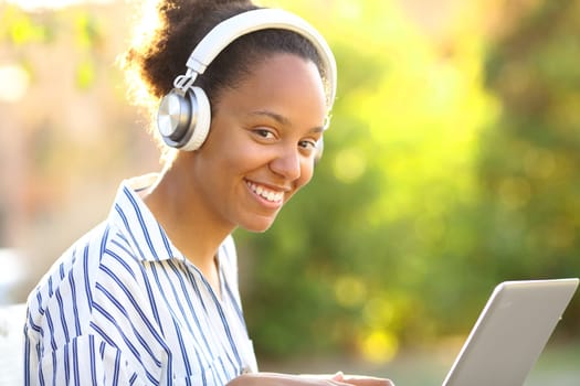 Happy black woman with laptop and headphone looking at you in a park