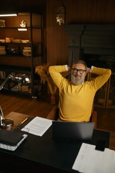 Excited mature man feeling satisfaction relaxed sitting at table with laptop in home office. Happy dreamy senior male holding hands behind head looking up with smile on face think of future success