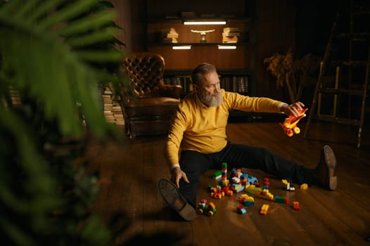 Happy enthusiastic old retired man playing toys while sitting on home floor over cozy eco-friendly interior design