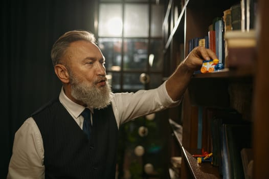 Senior businessman with old-fashioned outfit playing toy train on bookshelf in home or office library