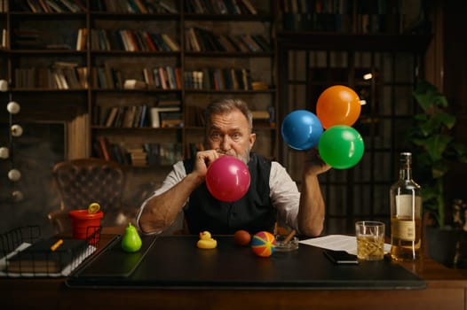 Portrait of senior business man blowing up balloon for success celebration. Grey-haired fashioned mature male boss sitting at work table in home office