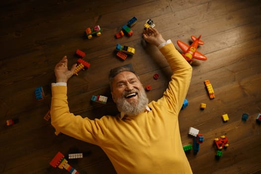 Happy senior man lying on floor with scattered toy building blocks. Overjoyed laughing elderly male pensioner celebrating success, having fun, dreaming about bright future on retirement. Overhead view