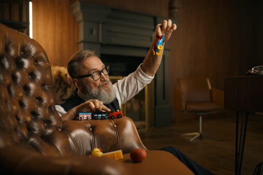 Funny senior businessman playing toys enjoying fun time after hard working day in home office. Elderly male sitting on floor nearby armchair