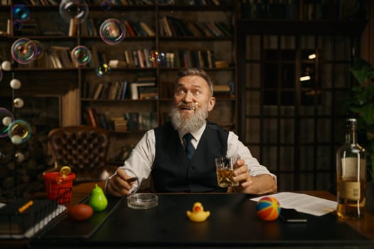 Portrait of relaxed senior businessman smoking cigar and drinking whiskey while sitting at work table in home office. Looking up at bubbles flying in air