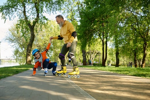 Senior man roller skating with little boy in urban park. Cheerful grandfather catching grandson falling upside down. Happy parenting and childhood