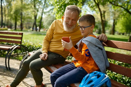 Grandfather and grandson watching video or taking selfie on smartphone while rest after roller skate riding in park. Happy family enjoying free time on weekend