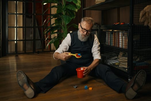 Funny senior businessman playing toy basket, scoop and building blocks while sitting on floor in luxury home office