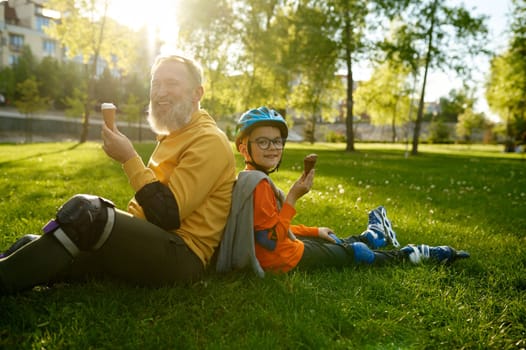Tired and happy grandfather and grandson having ice-cream after roller skating while sitting back to back on green grass street field