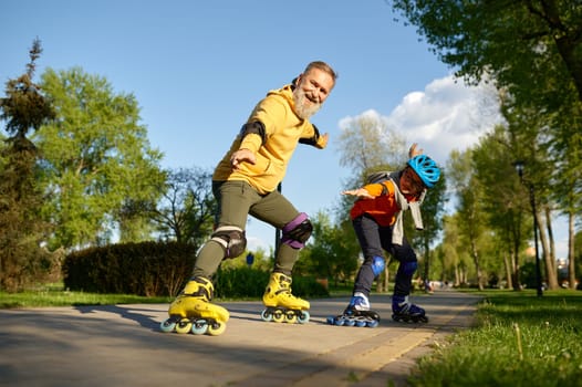 Happy grandfather and grandson on roller skates posing for camera over city park background. Playful weekend time and sports activity concept
