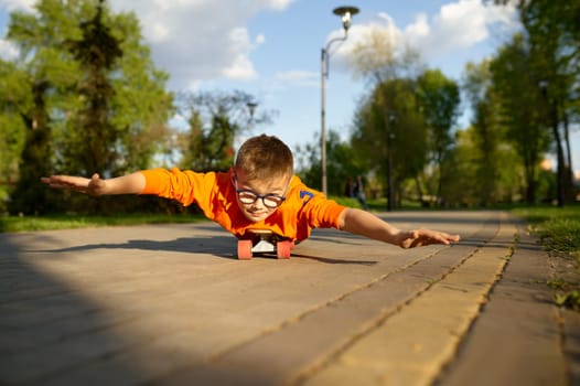 Boy lying on skateboard on his belly and riding like aviator pretending to fly in sunny park. Happy youngster enjoying activity lifestyle and extreme sport hobby