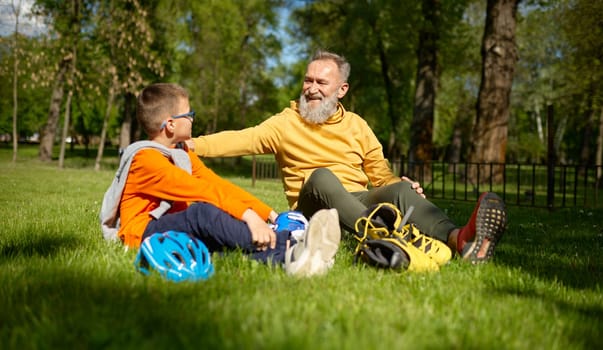 Positive smiling grandpa and grandson rest on green grass meadow in park. Granny supporting and praising grandchild for good riding roller-skates. Happy family recreation time on weekend