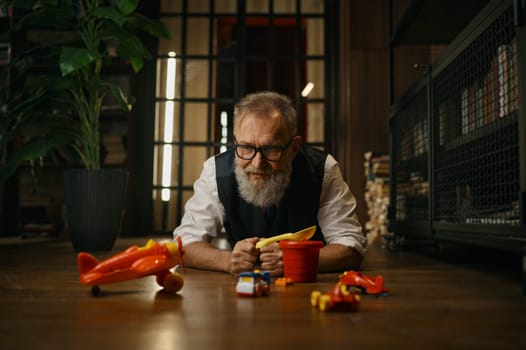 Funny successful senior businessman playing toys while lying on floor at home office. Relax time after work concept