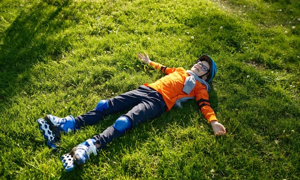 Happy tired little preschool boy wearing roller skates and helmet rest on green grass in urban park