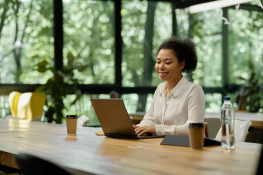 Satisfied smiling woman office worker looking at laptop screen typing message, communicating online or watching webinar. Good news, successful job and productive work concept