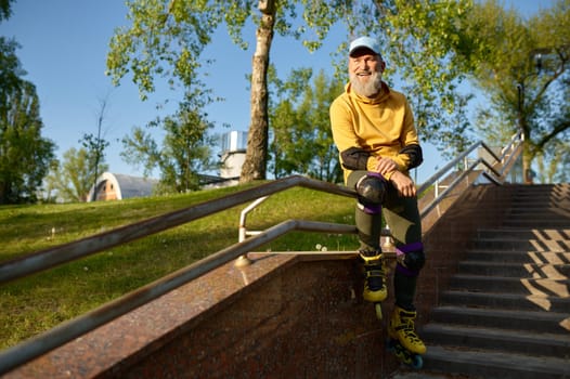 Happy smiling senior man wearing roller skates rest sitting on stair railing in urban park. Active time on retirement