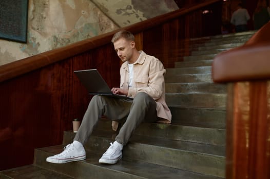 Man creative manager, online project developer or programmer working on wireless laptop computer sitting on stairs in office