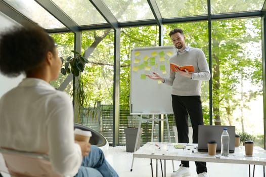 Man speaker showing notes on flipchart presenting new project ideas or marketing research results for colleague in eco-friendly boardroom