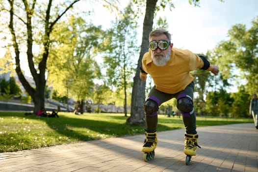 Senior man wearing funny goggles enjoying speed ride on roller skates over city park background at summer day