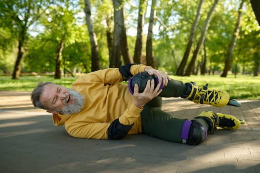 Senior man feeling pain in knee from injury falling down while roller skating in park. Accident and crash while rollerblading