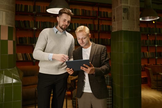 Two male business partners or web designers discussing project looking at mobile tablet while standing in office room of coworking space