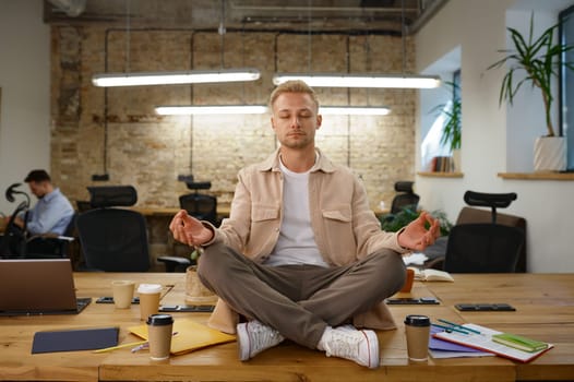 Calm man office worker meditating and breathing sitting in lotus position on desk table at workplace
