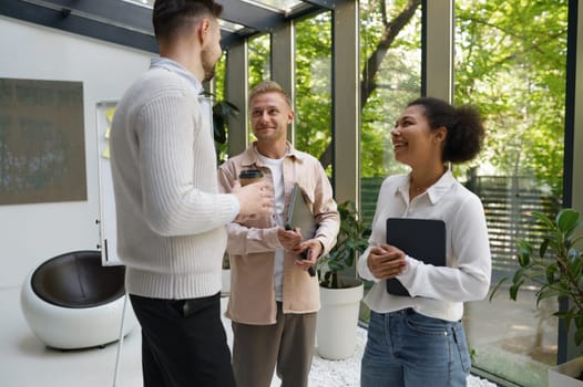 Smiling cheerful colleagues group standing at workplace and chatting. Happy positive businesspeople having fun conversation drinking coffee at work break