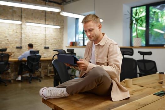 Confident concentrated man creative worker sitting on office table with digital tablet working with client online or developing new software design