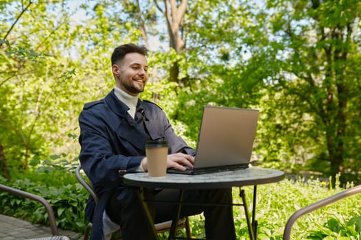 Businessman using laptop drinking coffee at open air cafe in park Independent male contractor working outdoors enjoying freelance and remote job