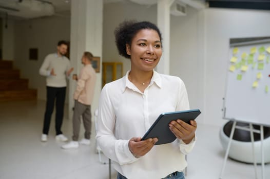 Portrait of female office worker holding digital tablet in hand looking forward while business colleagues talking on blurred background. Successful meeting and negotiation concept