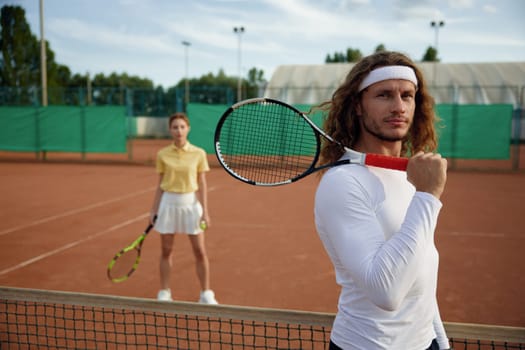 Friends playing tennis on court. Focus on confident man player holding racket on shoulder and looking in camera. Weekend sports recreation for young people couple
