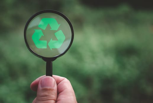 Concepts of waste reduction, pollution, reuse, efficient use of resources. Protect the environment by recycling. Human hand holding a magnifying glass with recycling sign on nature background.